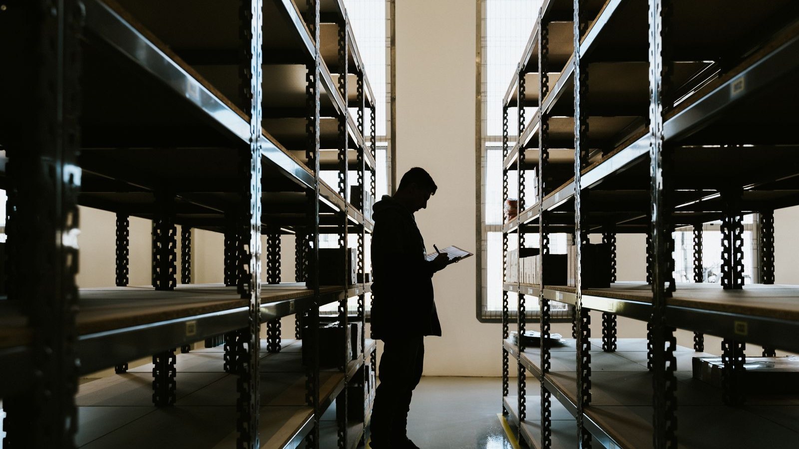 A worker in a high-visibility vest writing on a clipboard in a warehouse aisle